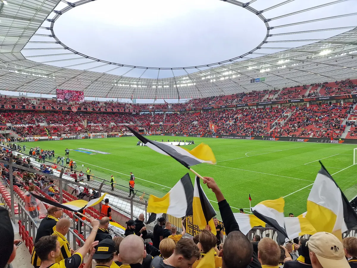 Inside the BayArena in Leverkusen before a match, with full red stands under the roof and visiting fans waving flags in the foreground