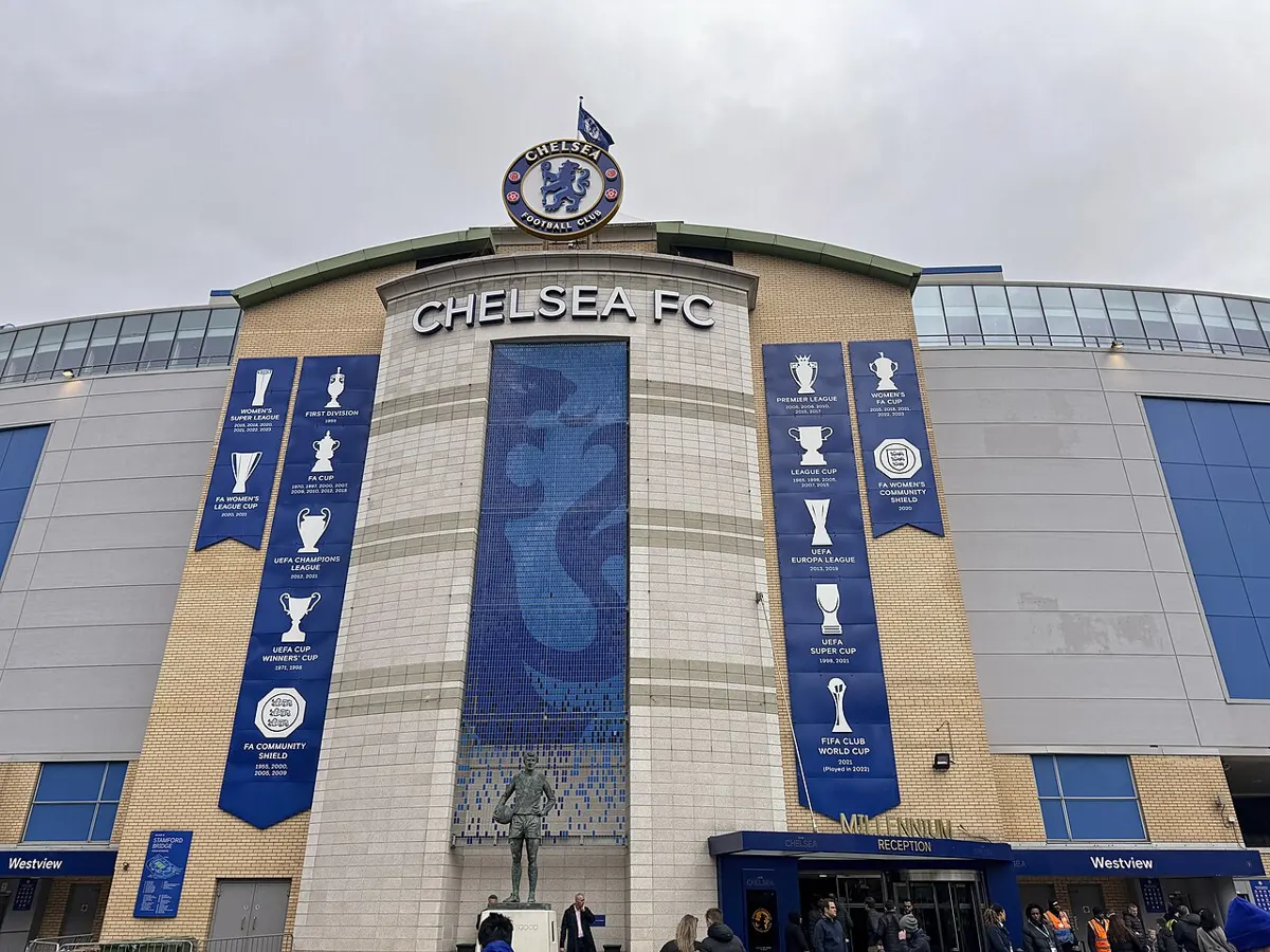 The Chelsea FC main entrance at Stamford Bridge, with the club crest above the stand, blue trophy banners on the wall and the Peter Osgood statue in front, in February 2024