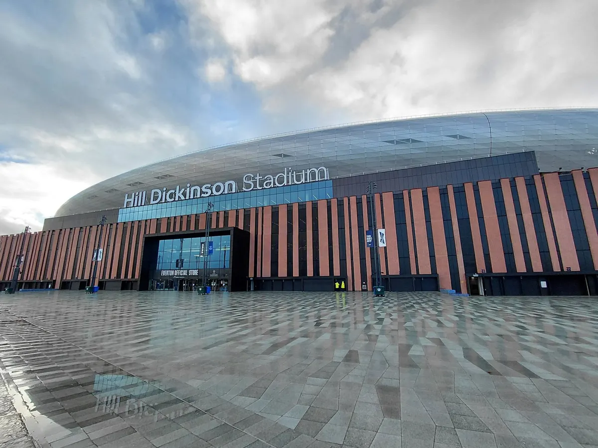 Exterior of Everton's Hill Dickinson Stadium at Bramley-Moore Dock in Liverpool, with the stadium name above the club store entrance under a cloudy sky