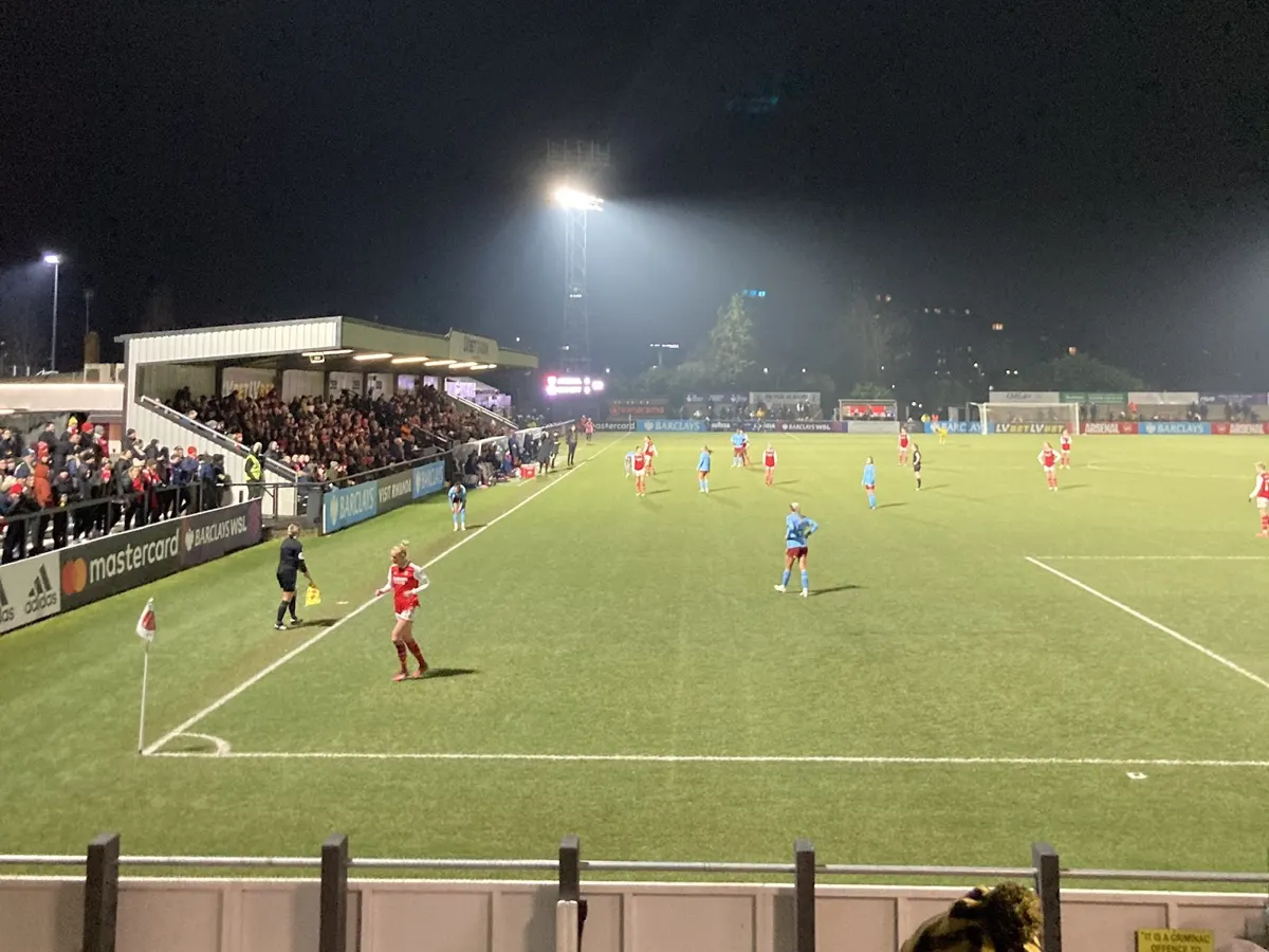 Arsenal Women players on the floodlit pitch at Meadow Park in Borehamwood during a 2023 match, with fans in the stand and along the touchline